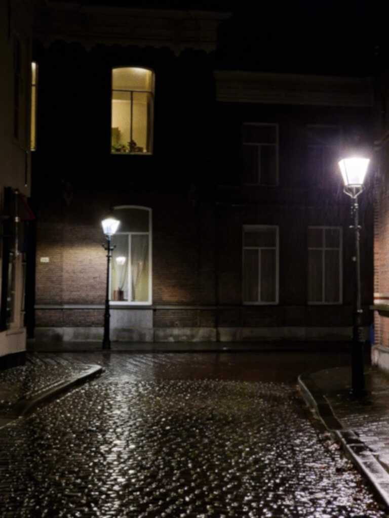 An empty, wet cobblestone street at night. It's lit by two lamp posts and a softly lit window in the dark brick buildings.